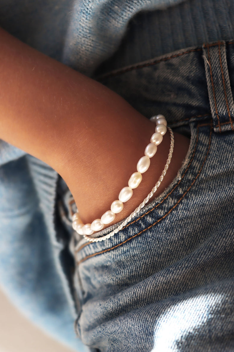 Pärlarmband med vita barockpärlor kombinerat med smalt silverarmband. Tjej som har handen nedstoppad i jeansfickan.