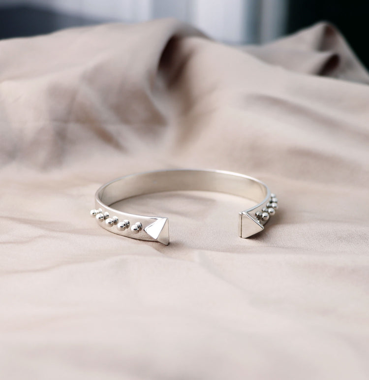Stelt silverarmband som ligger ner på ett tyg, armbandet har två stora pyramid-former och en rad av små silverkulor längs armbandet.