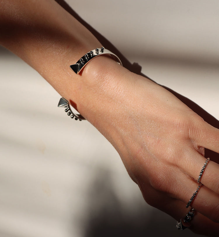 Tjej som bär ett stelt silverarmband med små silverkulor längs med armbandet och två stora nitar vid öppningen. Hon bär flera silverringar på olika fingrar med nitar.