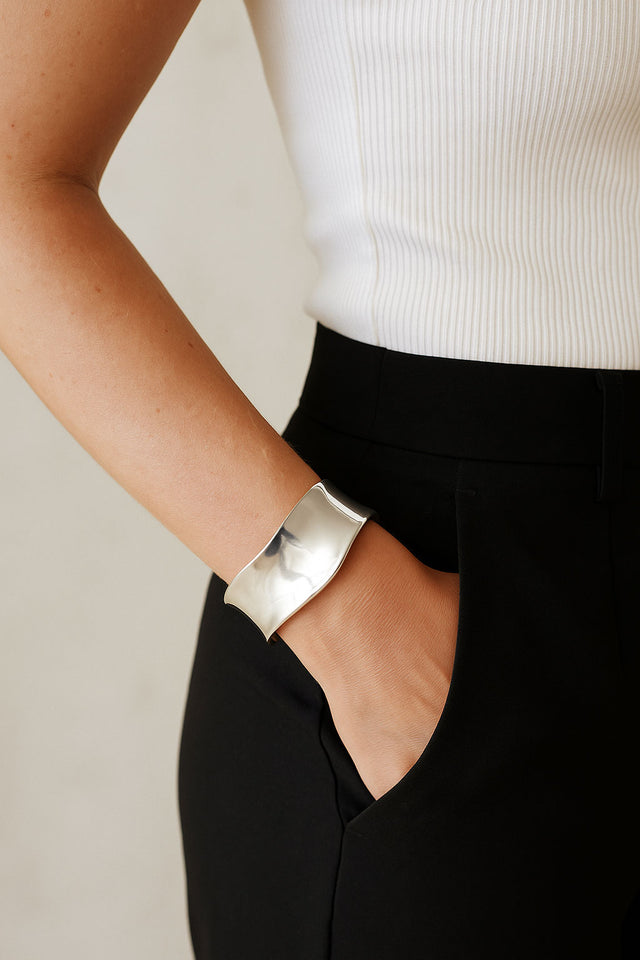 Kvinna som bär ett brett silverarmband med vågig form. Ett stilrent cuff armband i äkta silver. Hon bär svarta kostymbyxor och ett vitt ribbat linne.
