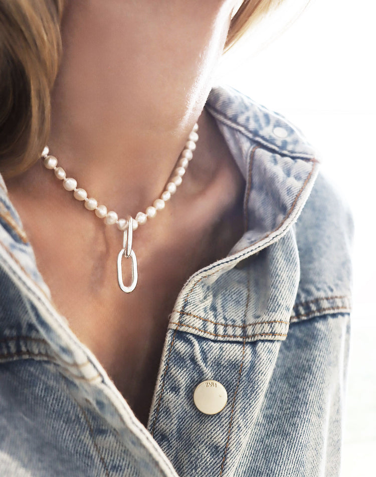 Tjej i jeansjacka som bär ett kort pärlhalsband som har två ovala silverlänkar som hängsmycke.