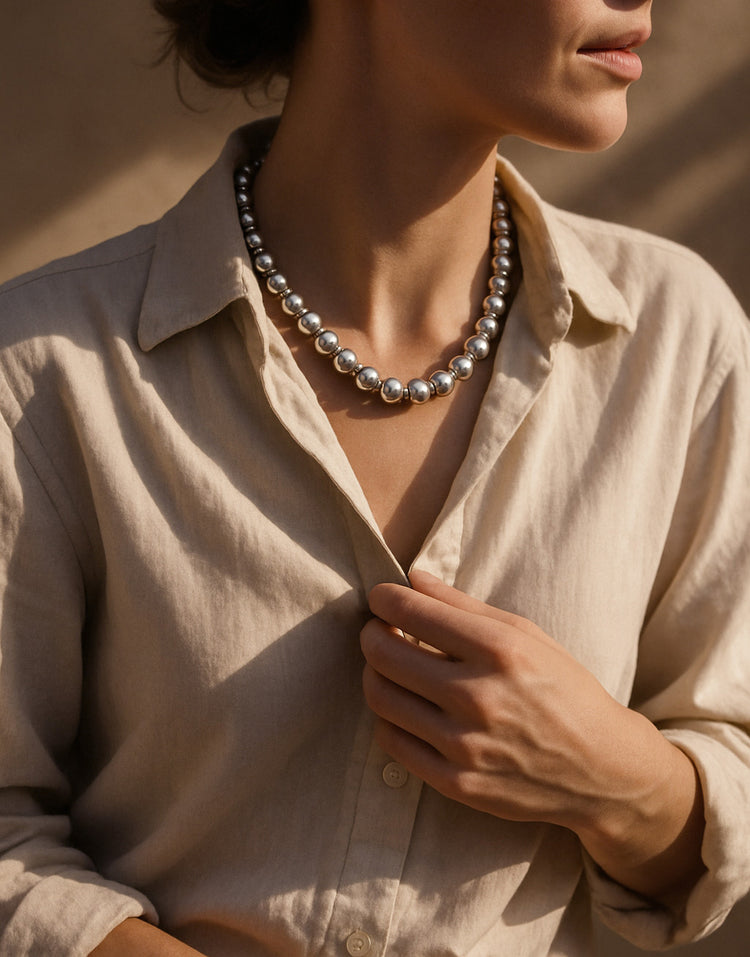 kvinna som bär ett stort silverhalsband med kulor. hon bär en beige linneskjorta och är utomhus med mjukt solljus.