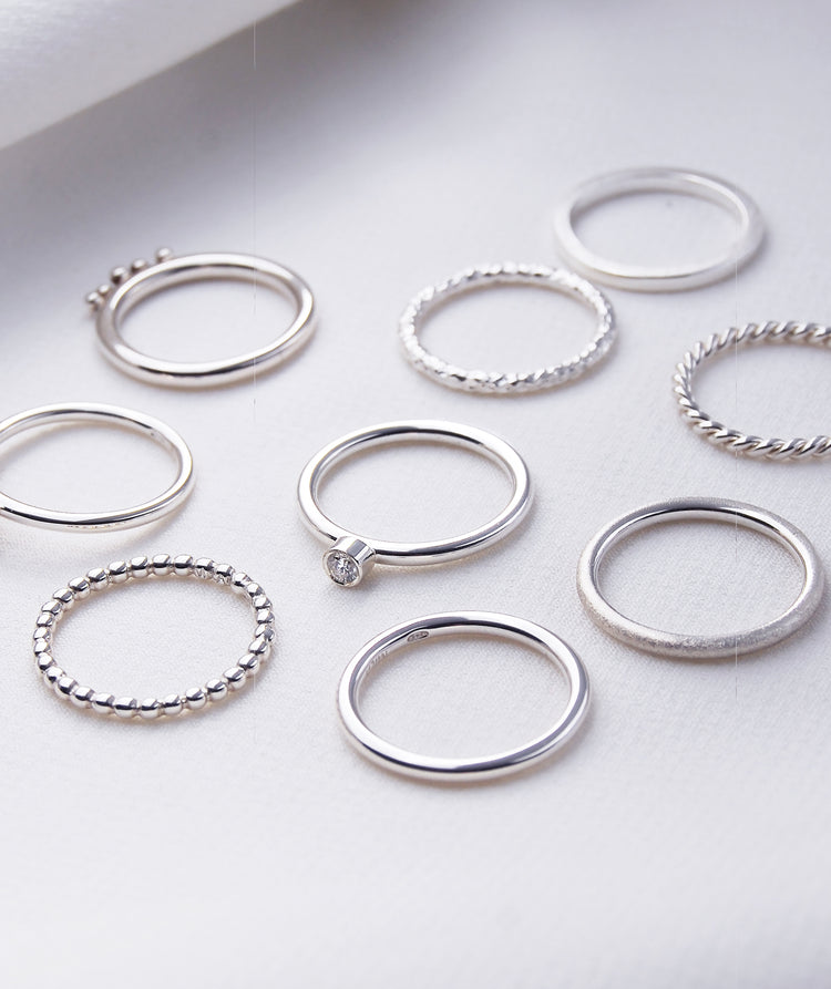 Många silverringar placerade på ett bord. Ringen i mitten har en diamant i bezel fattning.