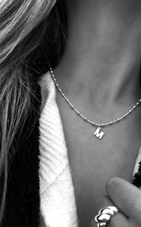 Tjej som bär ett bokstavshalsband i silver. Kedjan har små kulor med berlock - bokstav M.