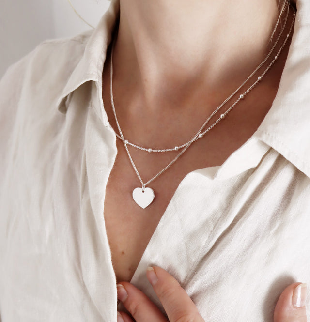 En kvinna som bär två silverhalsband. Ett kortare med kedja och silverkulor och ett längre med ett blankt silverhjärta i kedja. 