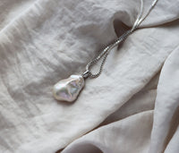 Halsband med stor pärla som har ojämn form placerad på ett mjukt tyg. 