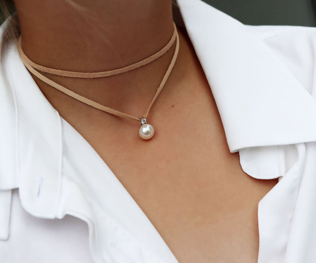 Tjej som bär ett kort pärlhalsband med naturfärgat läderband virat runt halsen och pärlberlock som är utsmyckad med en gnistrande diamant. Hon bär vit uppknäppt skjorta. 