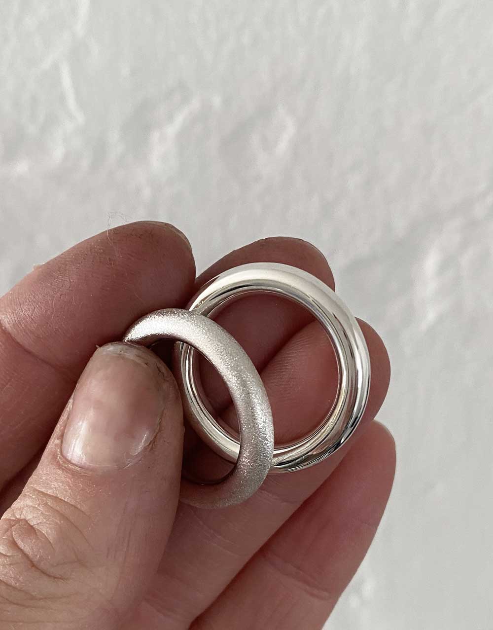 två ringar i silver som hålls mot varandra. En med mattslipad yta och en ring med blank, slät yta. 